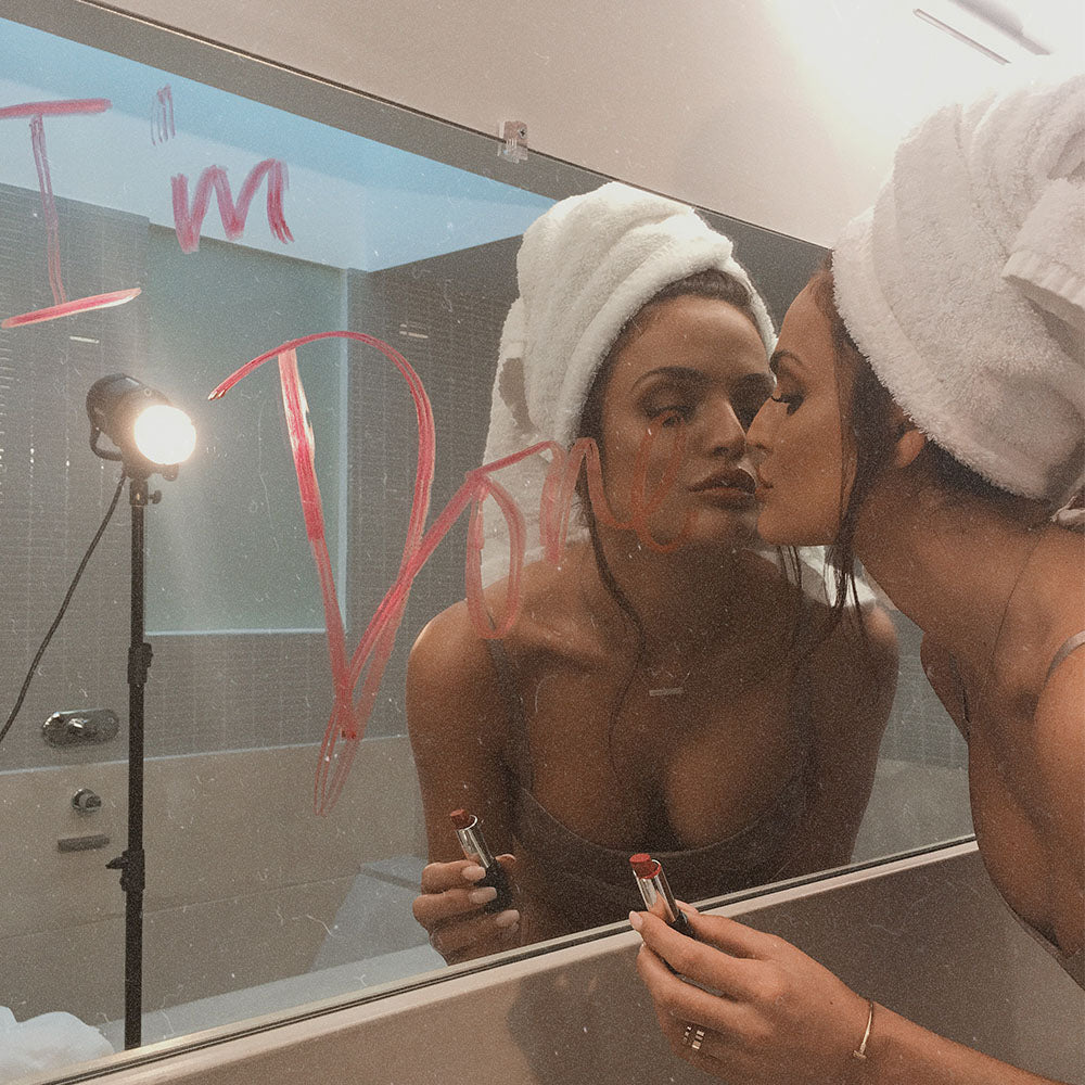 Woman with a towel wrapped on her head applies lipstick in a bathroom mirror while her reflection leans in as if kissing