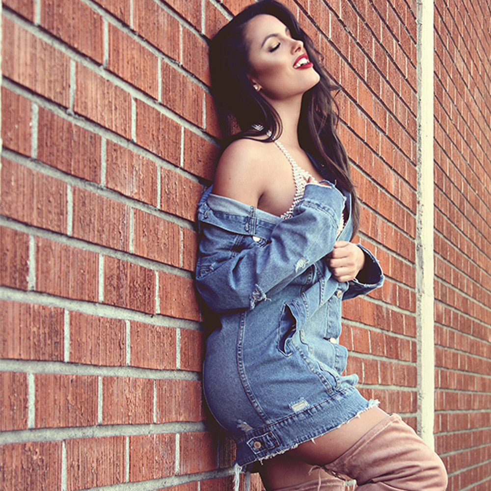 Confident model in an off shoulder denim jacket and short distressed dress leans against a brick wall and smiles.