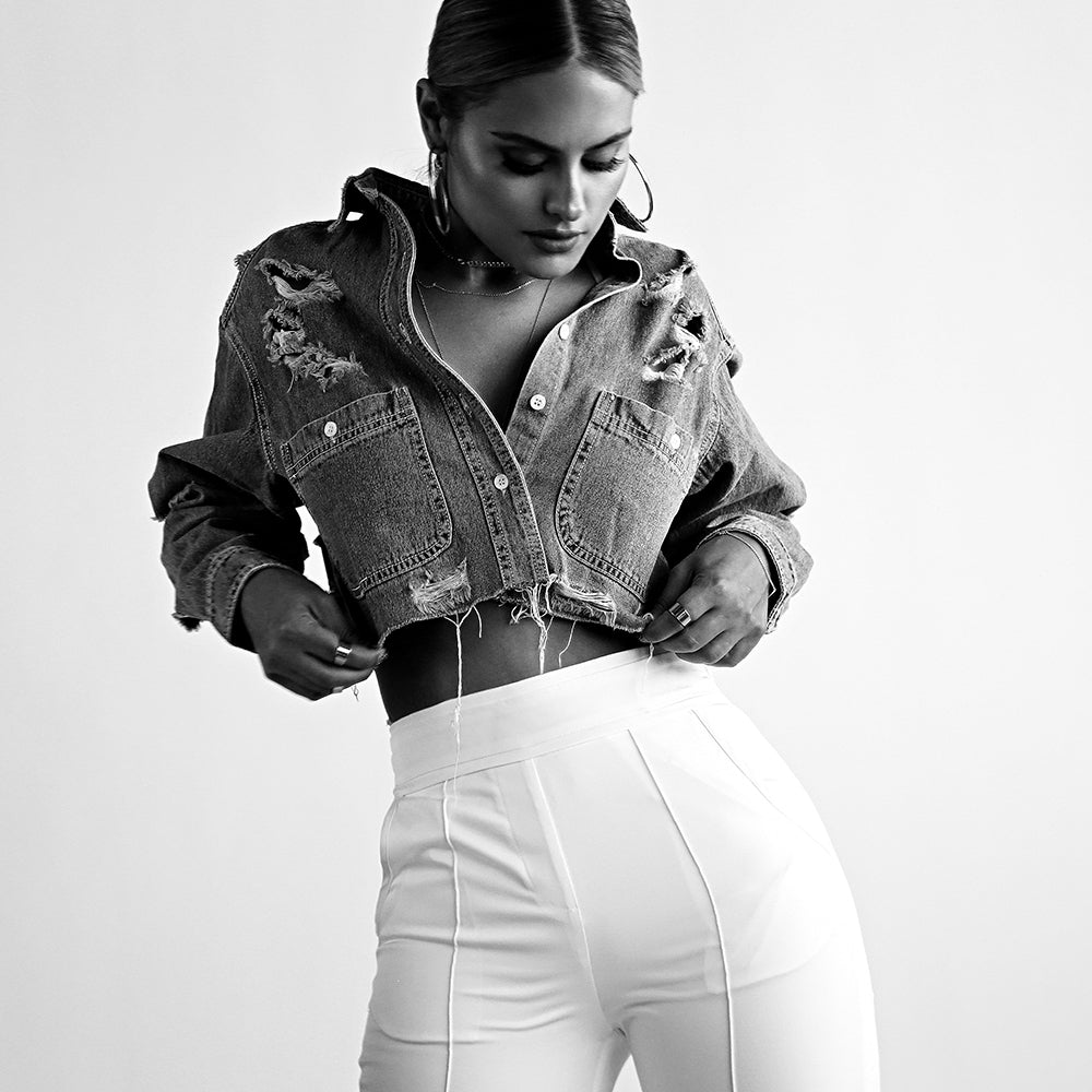 Black and white close-up of a model wearing a cropped distressed denim jacket and high-waisted white pants.