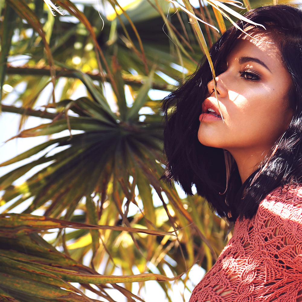 Woman with dark hair in a pink lace top posing among tropical palm leaves in bright sunlight.