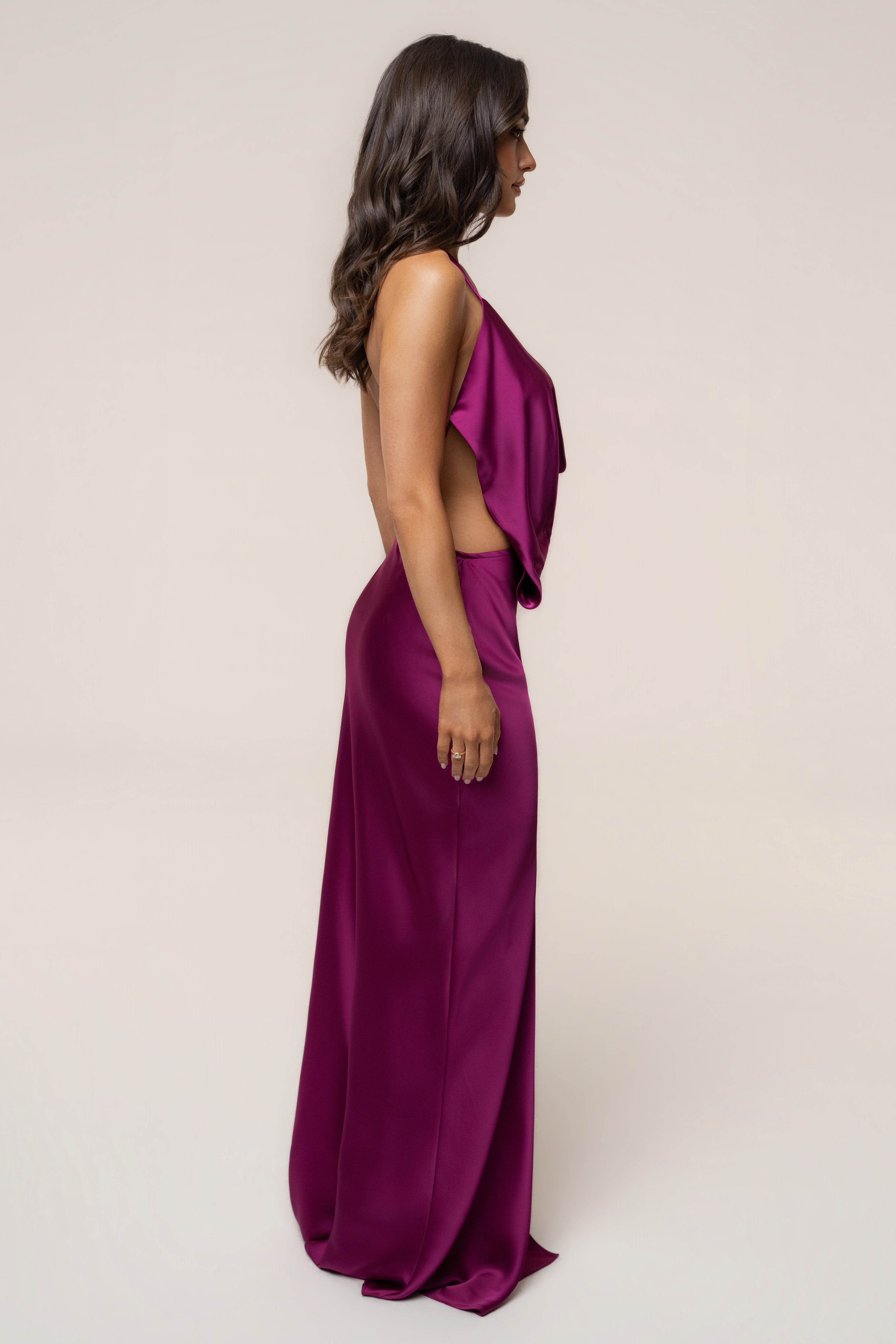 Side profile of a model wearing a fuchsia Delmara Cowl Maxi Dress with open back and a long flowing skirt