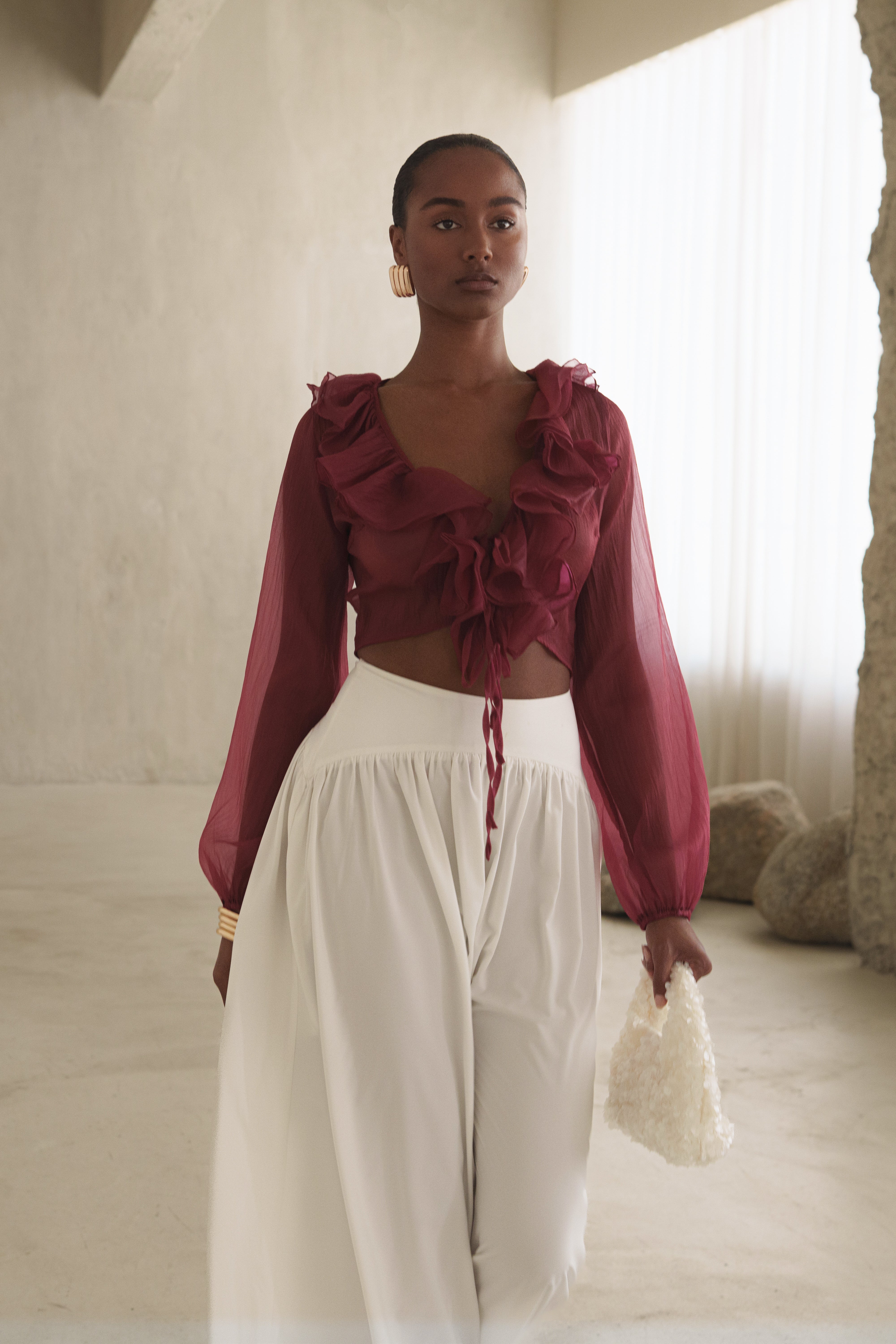 Full length side view of model in berry Delilah ruffled top with white high waisted pants and a cream bag