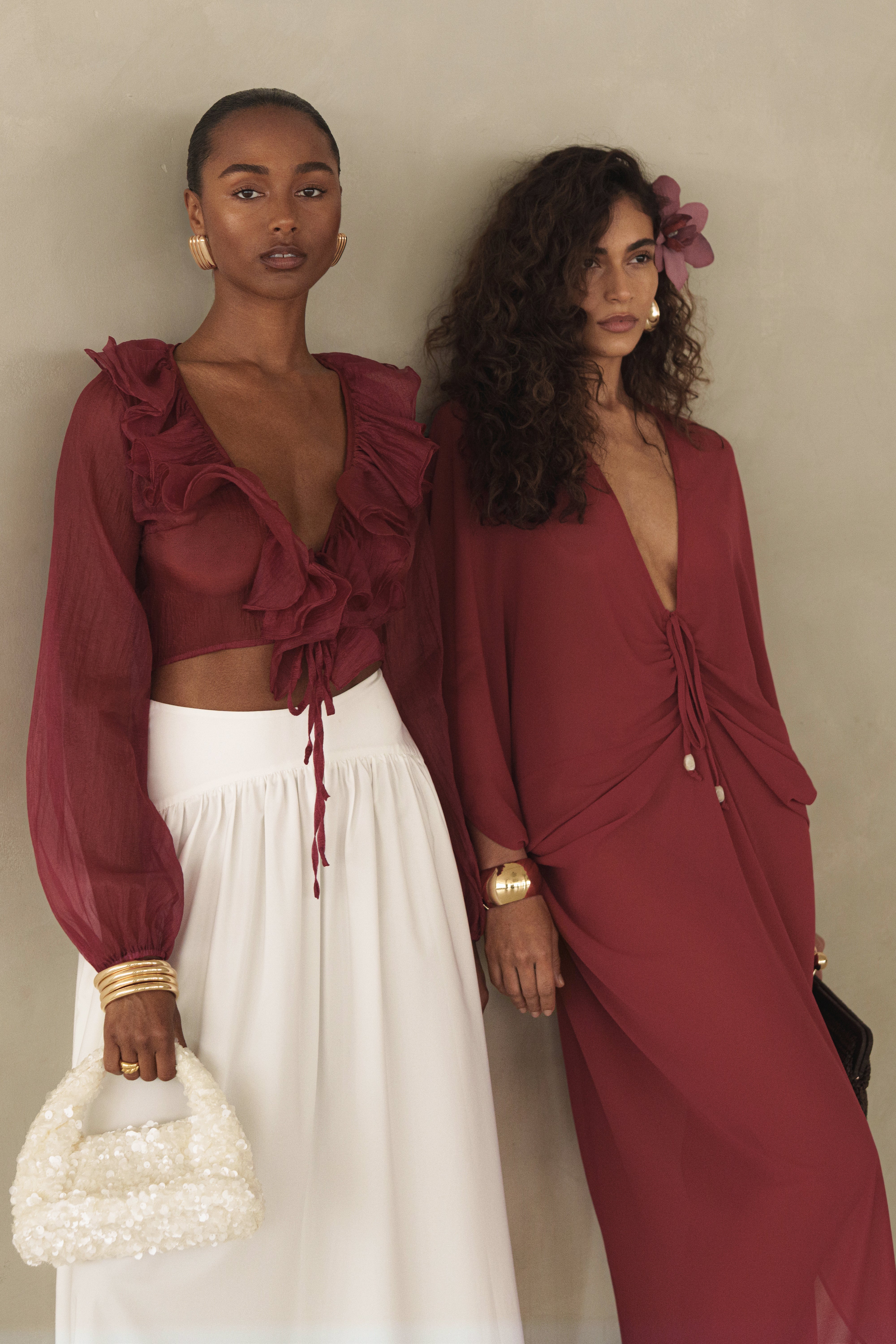 Two models wear Berry Delilah Ruffled Top looks in burgundy; left in a cropped ruffled blouse with sheer sleeves and white skirt, right in a burgundy wrap dress.