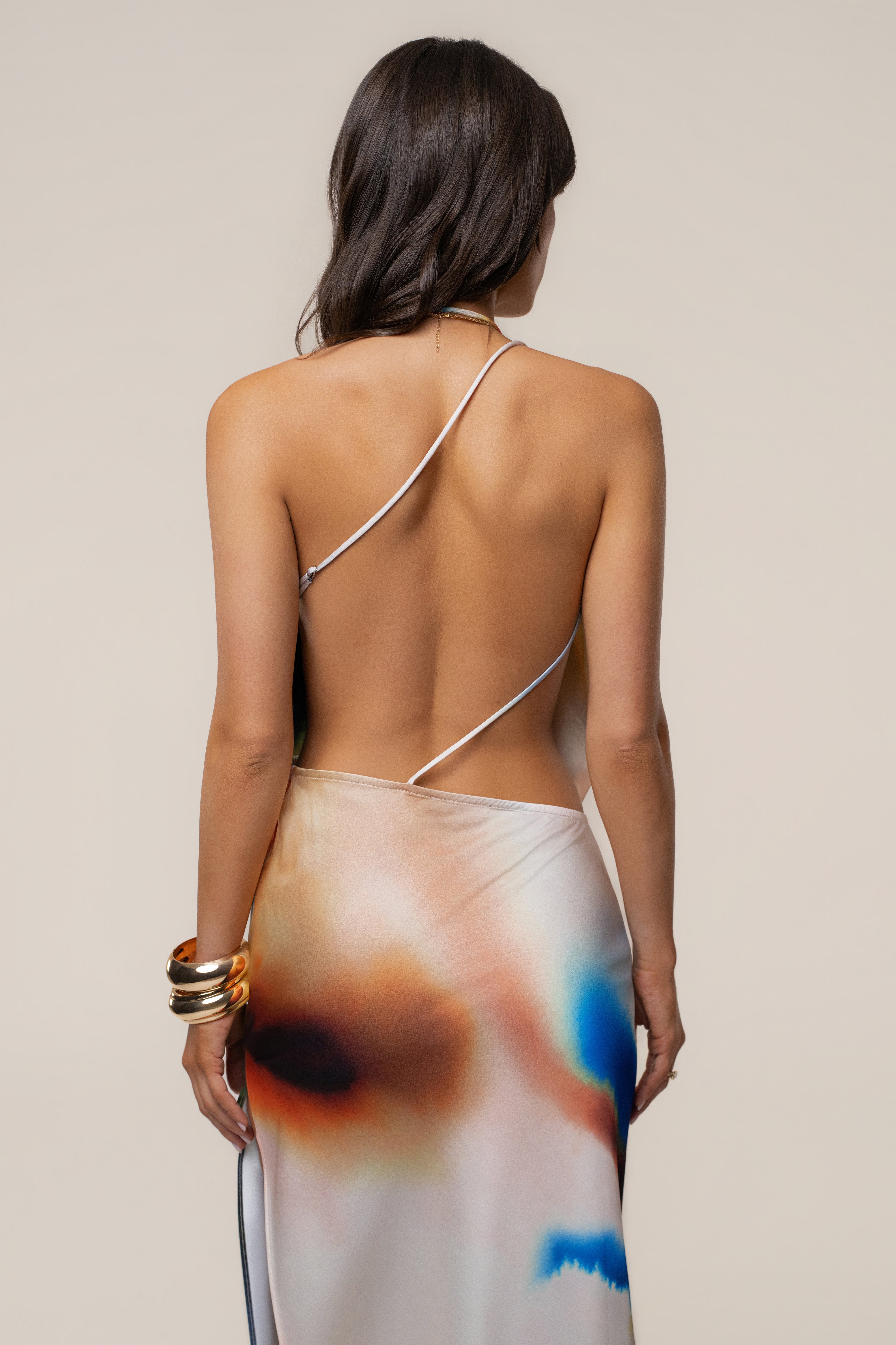 Back view of a woman wearing a white crossback maxi dress with a diagonal strap and a multicolor abstract hem