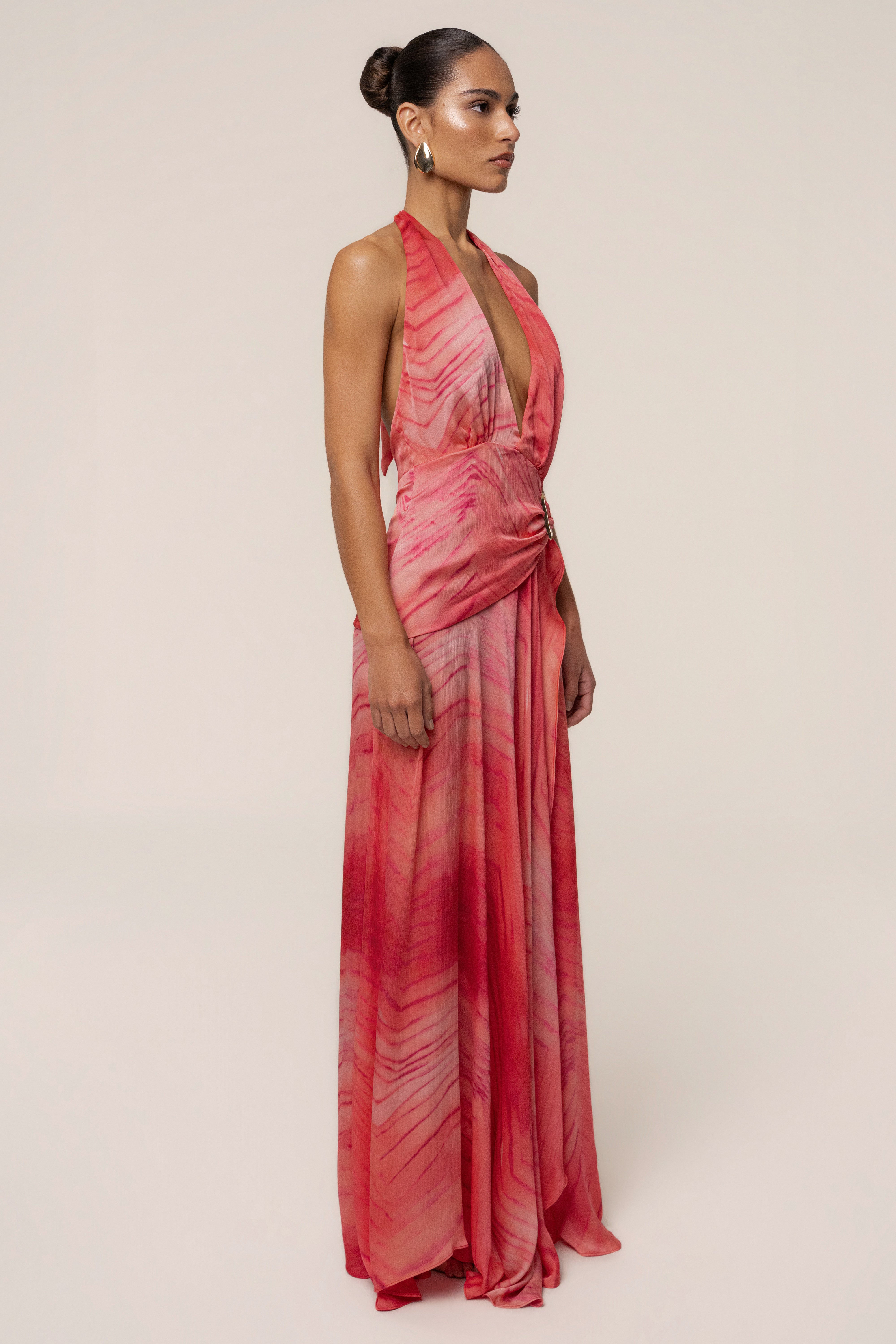 Side view of a model in coral red Vacation State maxi dress with halter neck open back and gold ring belt.